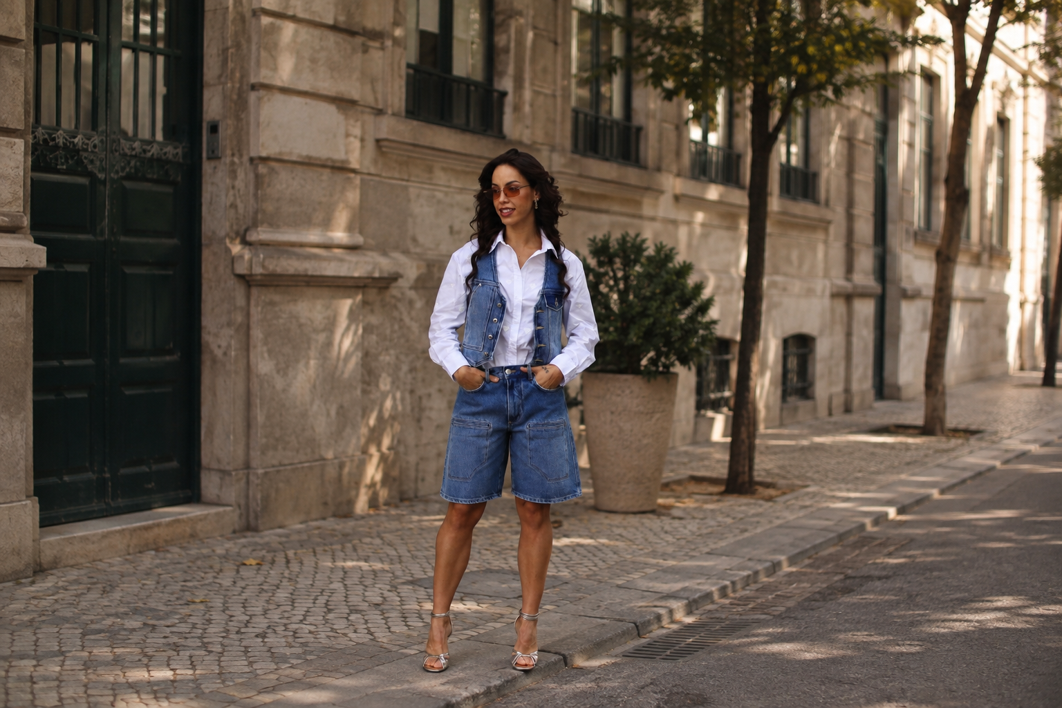 Mulher com look de transição verão-outono numa rua do Porto. Camisa branca, colete e calções de ganga e saltos altos dourados.